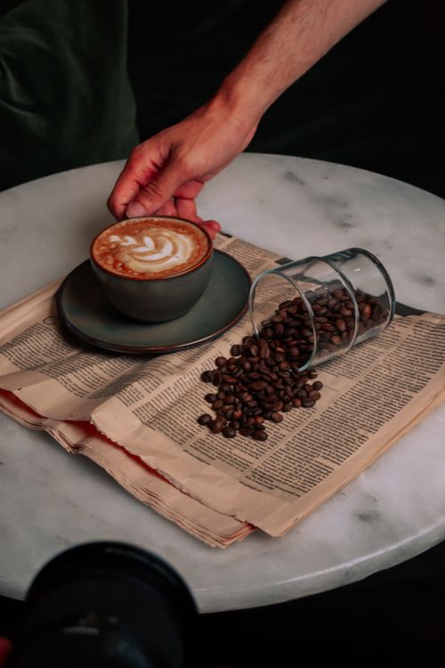 Free A hand reaching a latte with elegant latte art beside coffee beans on a paper. Stock Photo
