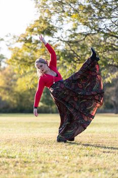 Woman in a vibrant dress performs a dance move in a sunny autumn park.