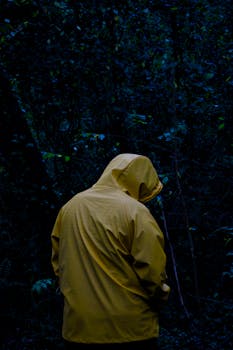 An individual in a yellow raincoat stands alone in a dense, dark forest, conveying solitude and mystery.