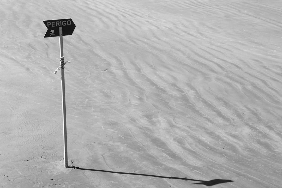 Black and white photo of a 'Perigo' caution sign on a sandy beach in Mongaguá, Brazil.