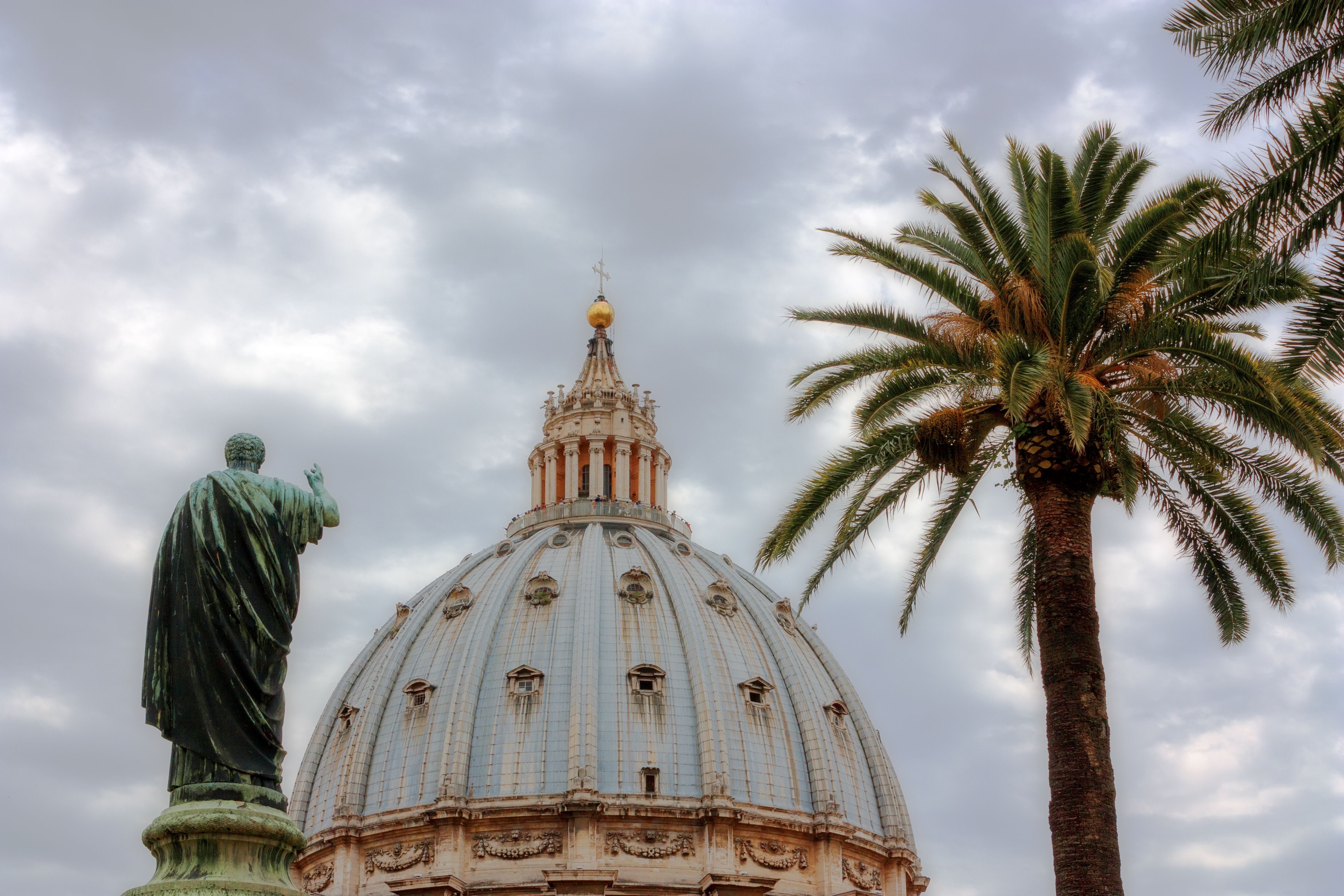 View of Saint Peter's Basilica dome with a sculpture and palm tree in Vatican City.