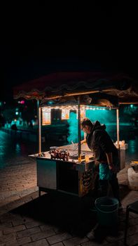 A street food vendor operating a brightly lit cart at night in an urban setting.
