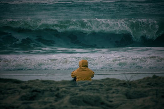A lone person in a yellow jacket sits on the beach, gazing at the powerful Atlantic Ocean waves in Nouvelle-Aquitaine, France.