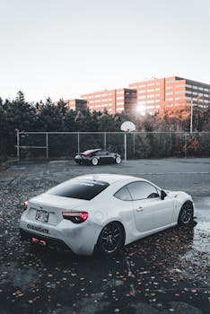 Two sports cars parked near an urban basketball court in autumn, trees and buildings in the background.