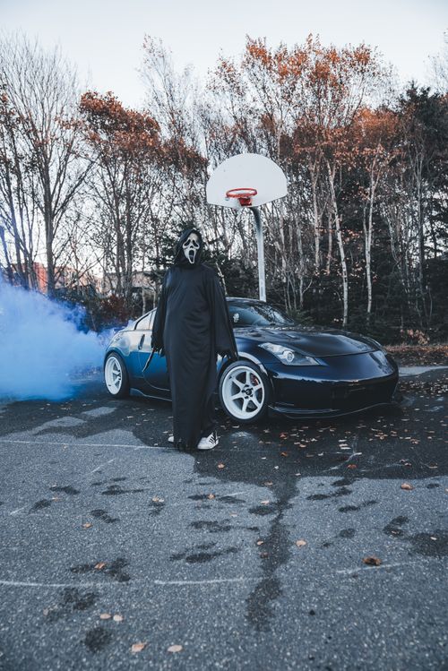 Free Halloween-themed scene with a masked figure in front of a sports car and smoke on a fall day. Stock Photo