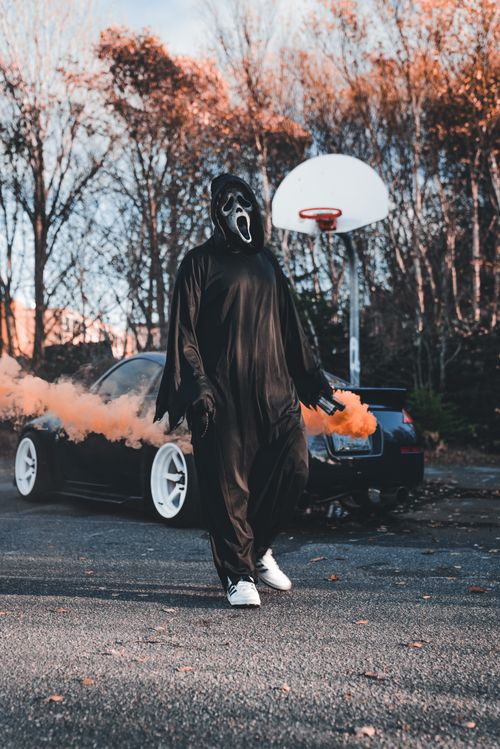 Free A masked figure in a black costume walks past smoke and a car, set against autumn foliage. Stock Photo