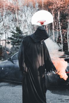 Mysterious hooded figure holding a smoke bomb near a car with backdrop of trees and basketball hoop.