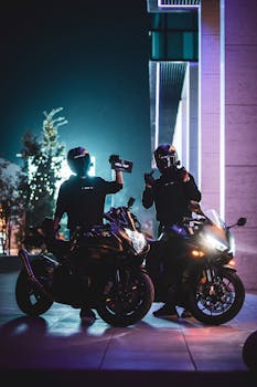 Two motorcyclists with helmets pose at night against a vibrant urban backdrop.