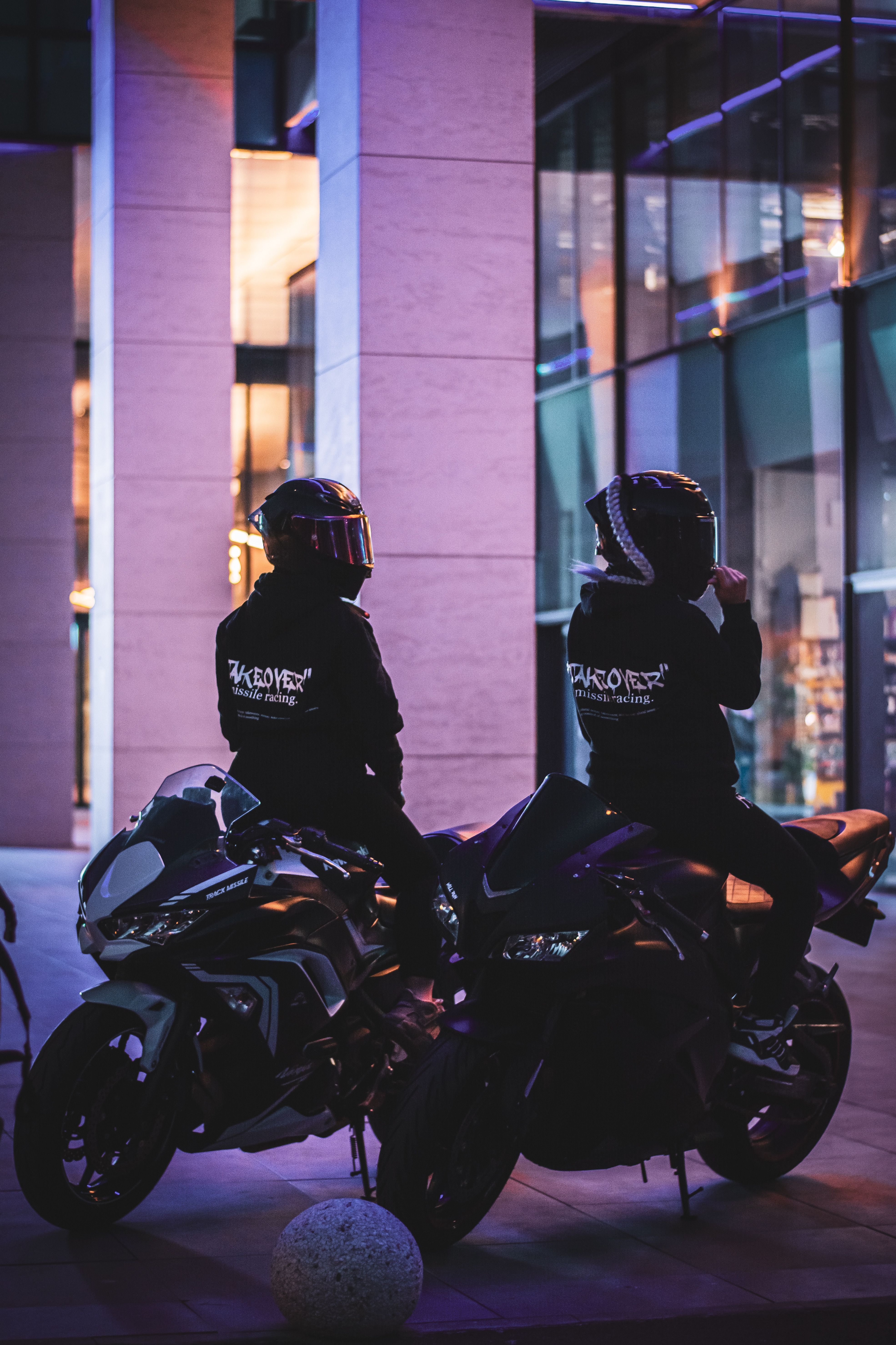 Two motorcyclists in helmets sitting on bikes in a night-time urban setting.