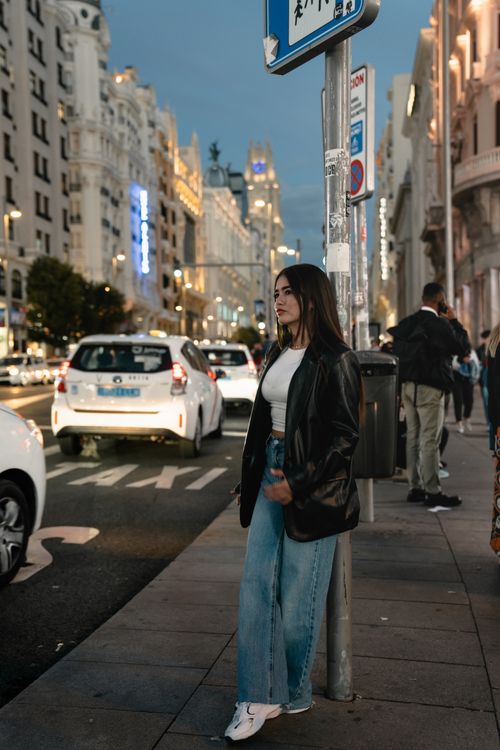 Free Woman in leather jacket and jeans stands on bustling city street at dusk, surrounded by cars and iconic architecture. Stock Photo