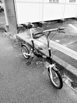 A black and white photo of a folding bike parked on a roadside in an urban setting.