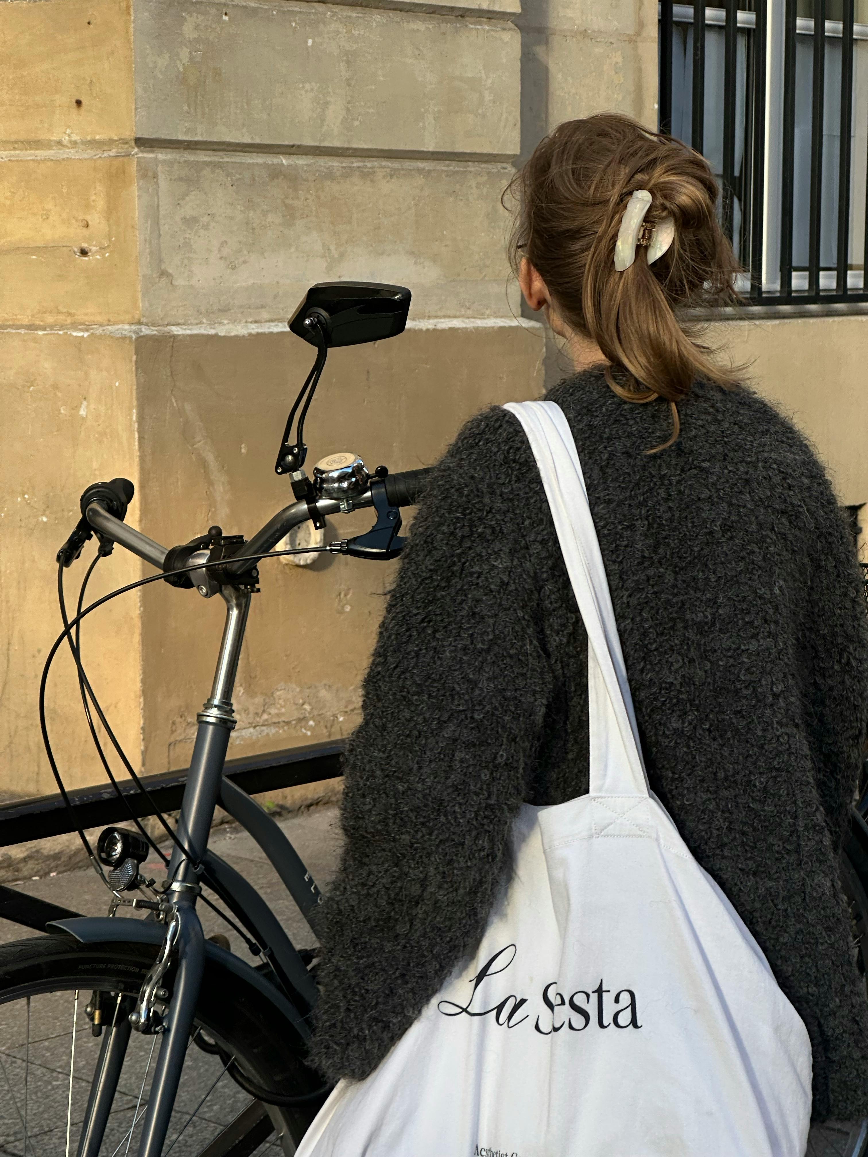 A woman with a bicycle standing by a stone wall, holding a tote bag.