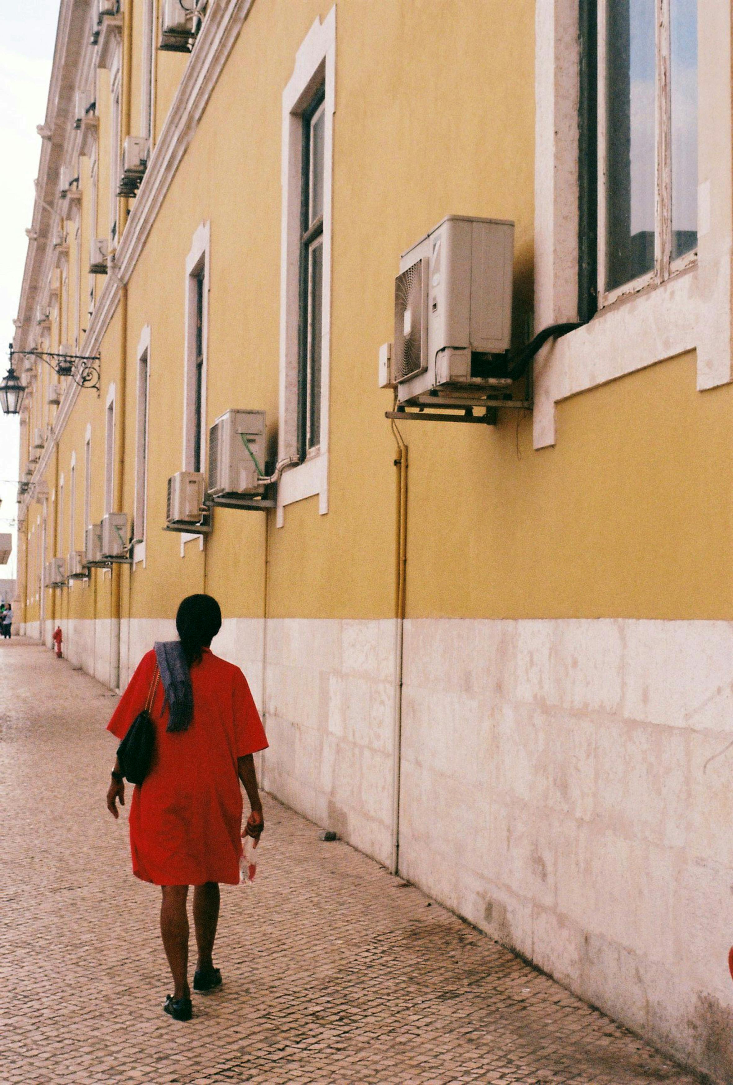 A person in a red dress walks beside a yellow historic building in Lisbon.