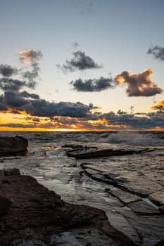 Stunning sunset over a rocky shoreline with vibrant clouds and waves crashing.