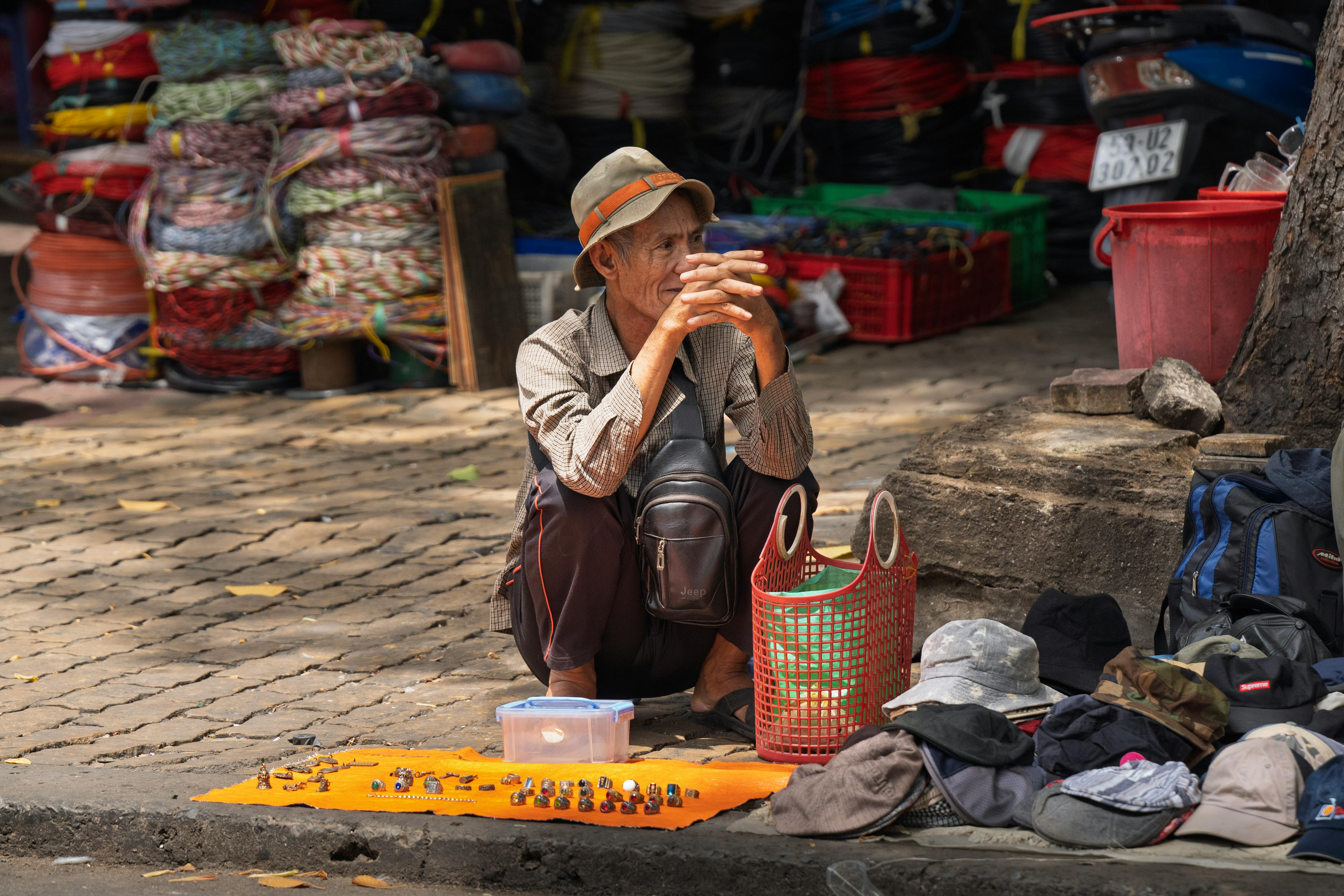 An elderly street vendor selling goods on a sunlit urban street corner.