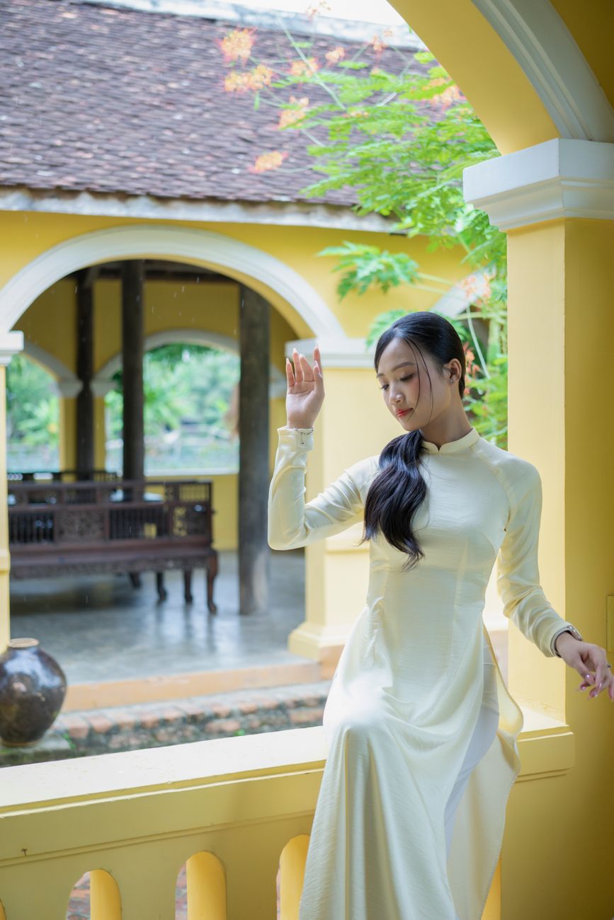Woman in white áo dài posing gracefully by a traditional Vietnamese structure.
