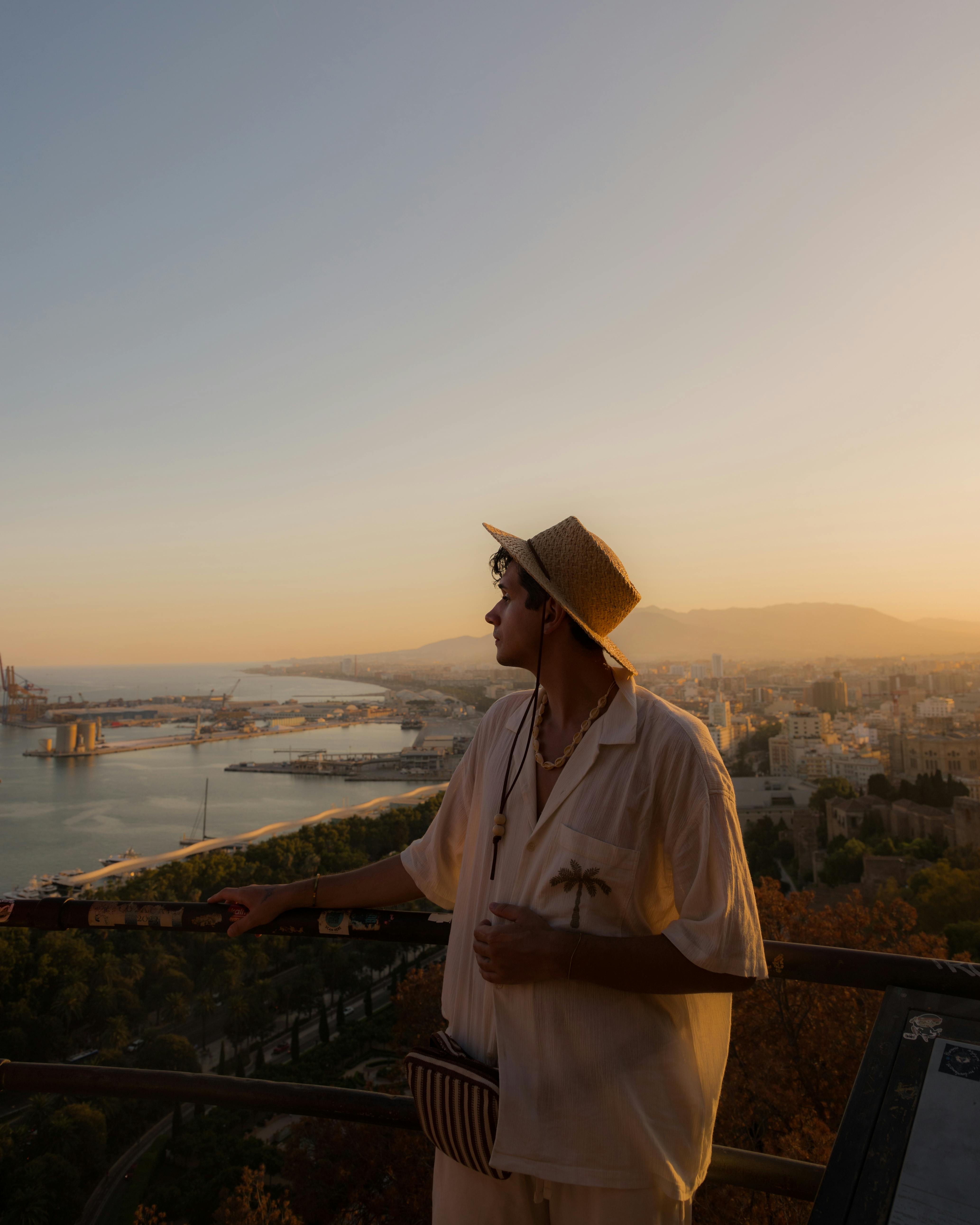 Man in stylish attire gazes over Málaga cityscape at sunset.