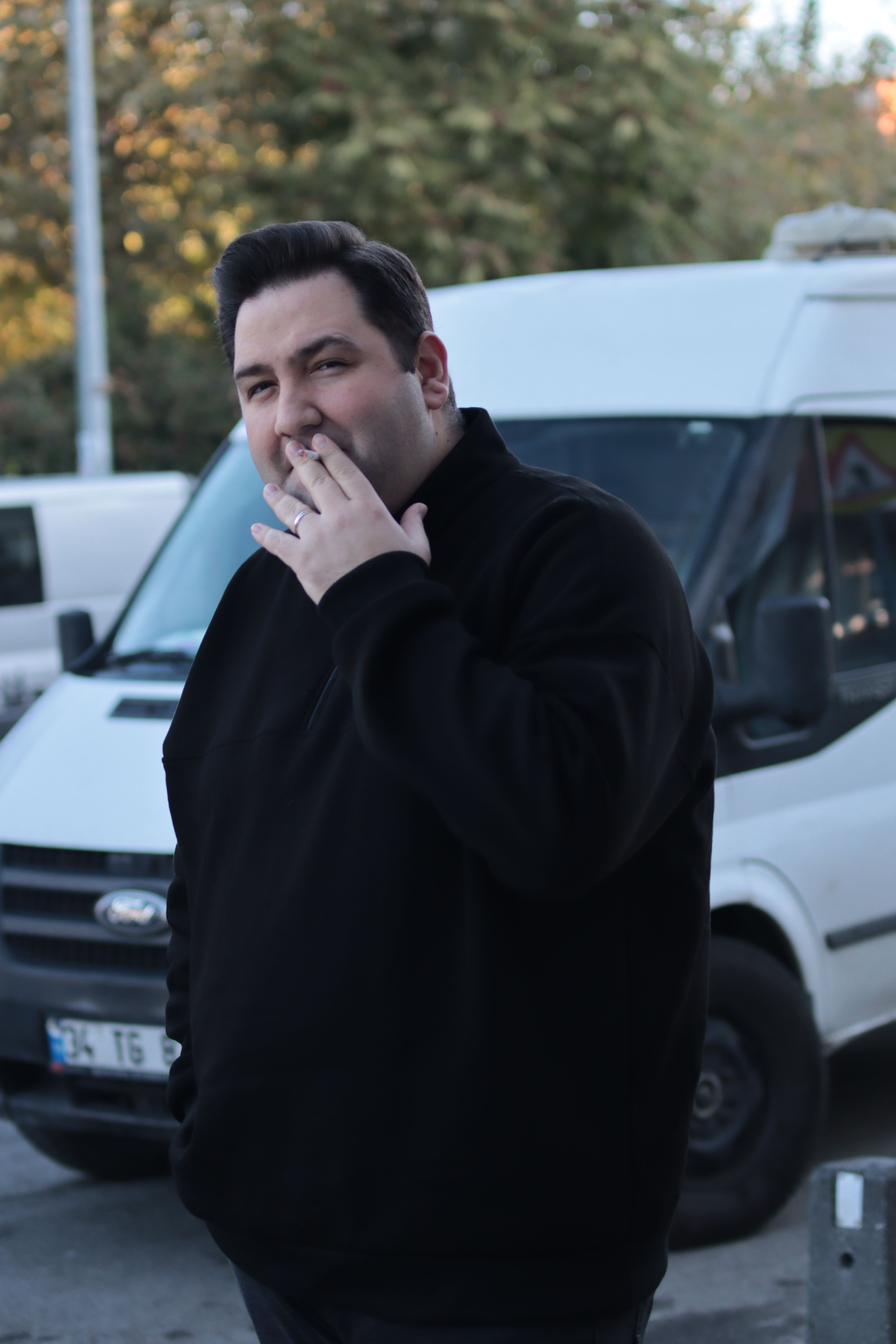 Adult man smoking outdoors beside a white vehicle in an urban setting.