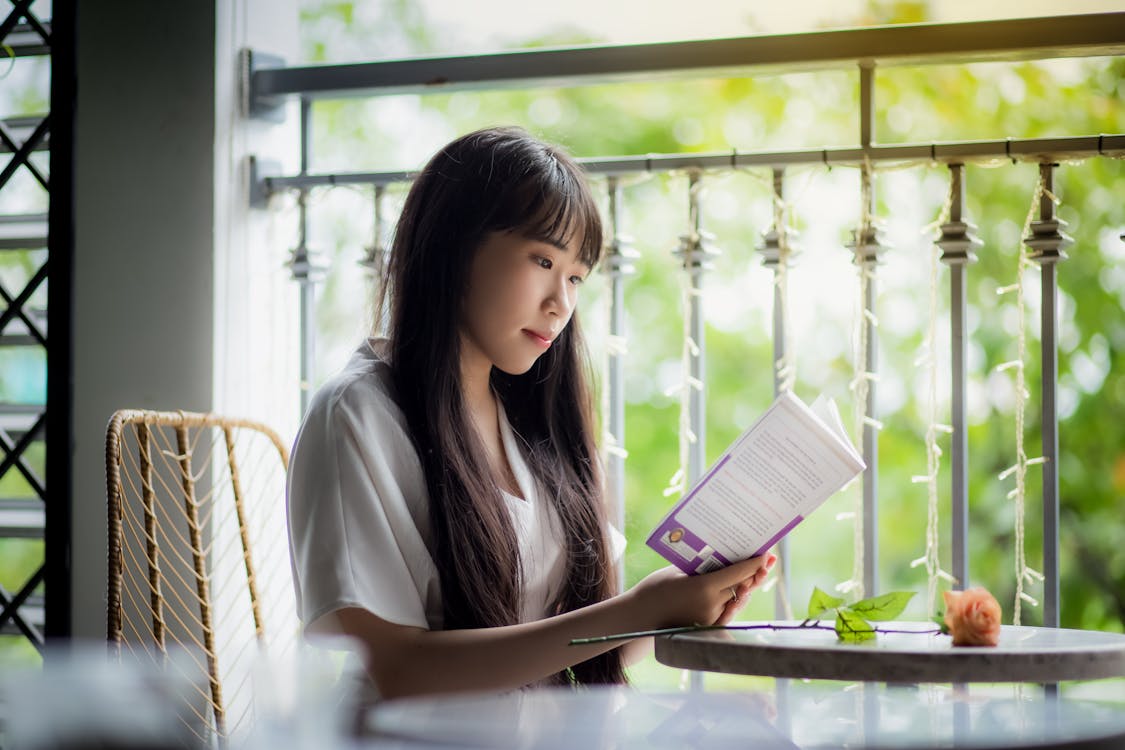 Free Woman Sitting on Chair Beside Table Reading a Book Stock Photo
