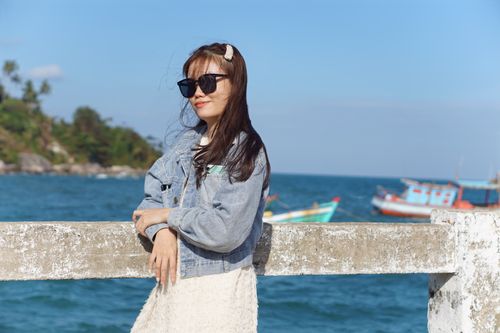 Free A young woman in sunglasses and denim jacket enjoying a sunny day by the ocean. Stock Photo