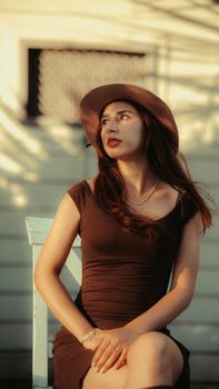 Elegant woman in hat sitting outside, bathed in warm sunset light with a fashionable look.