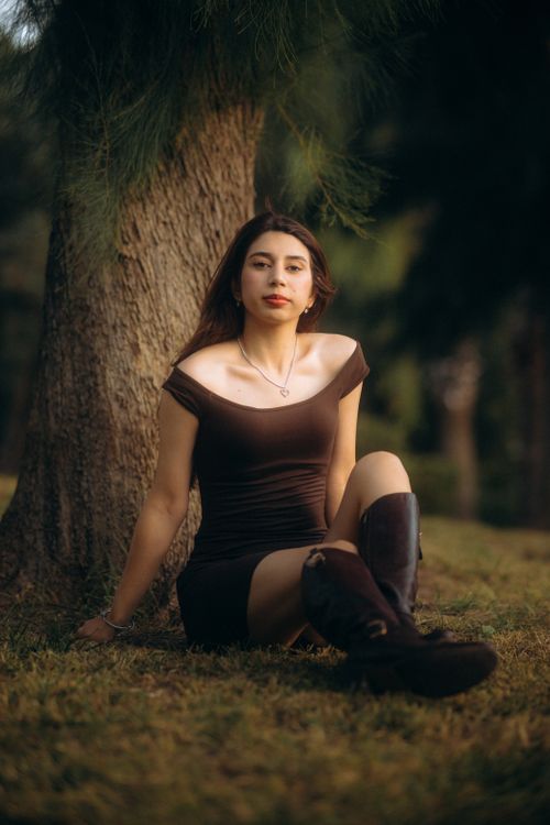 Free A young woman in a brown dress sitting by a tree in a tranquil autumn park. Stock Photo