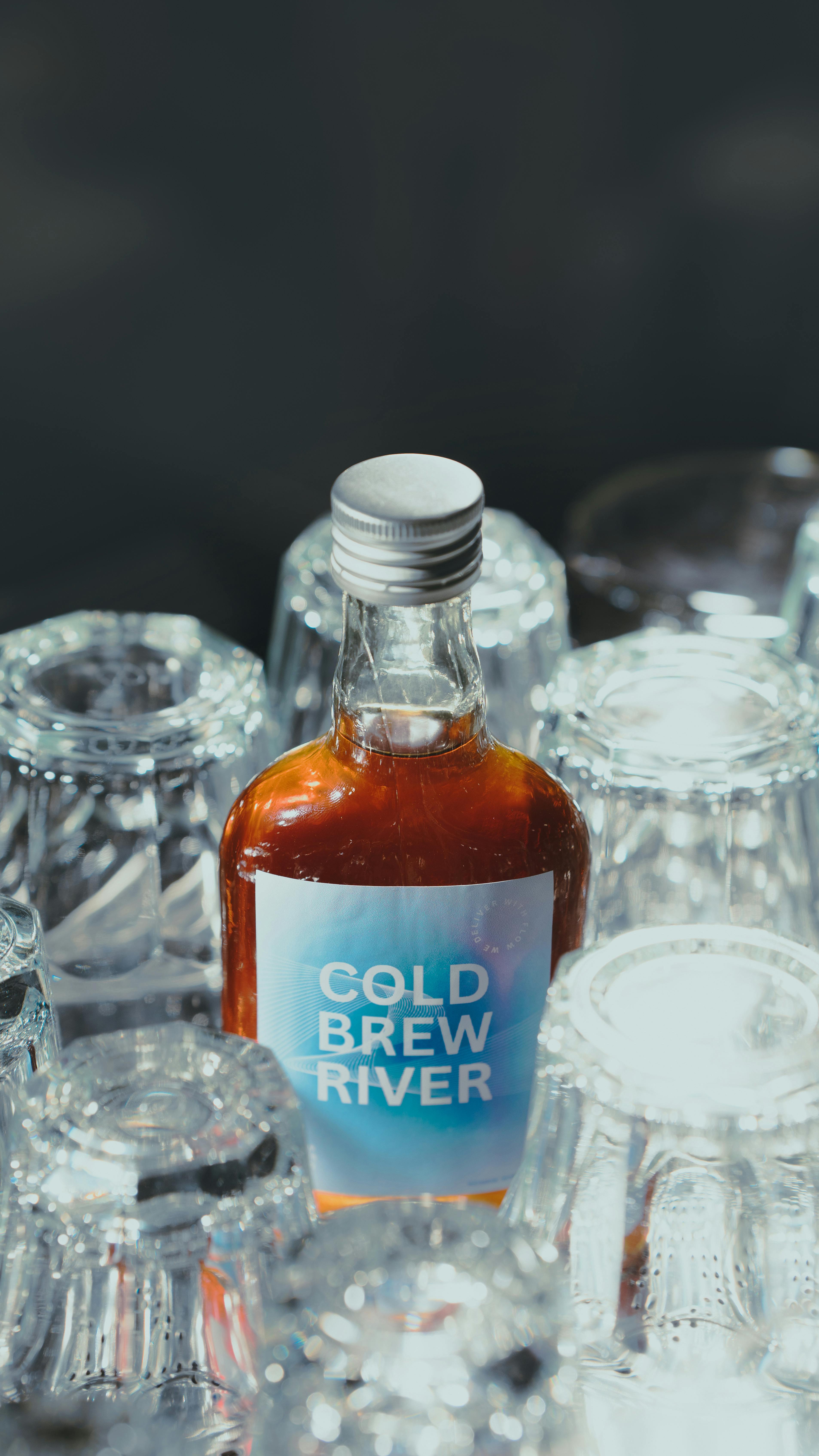 A cold brew coffee bottle placed among several upside down glasses, sunlight casting reflections.