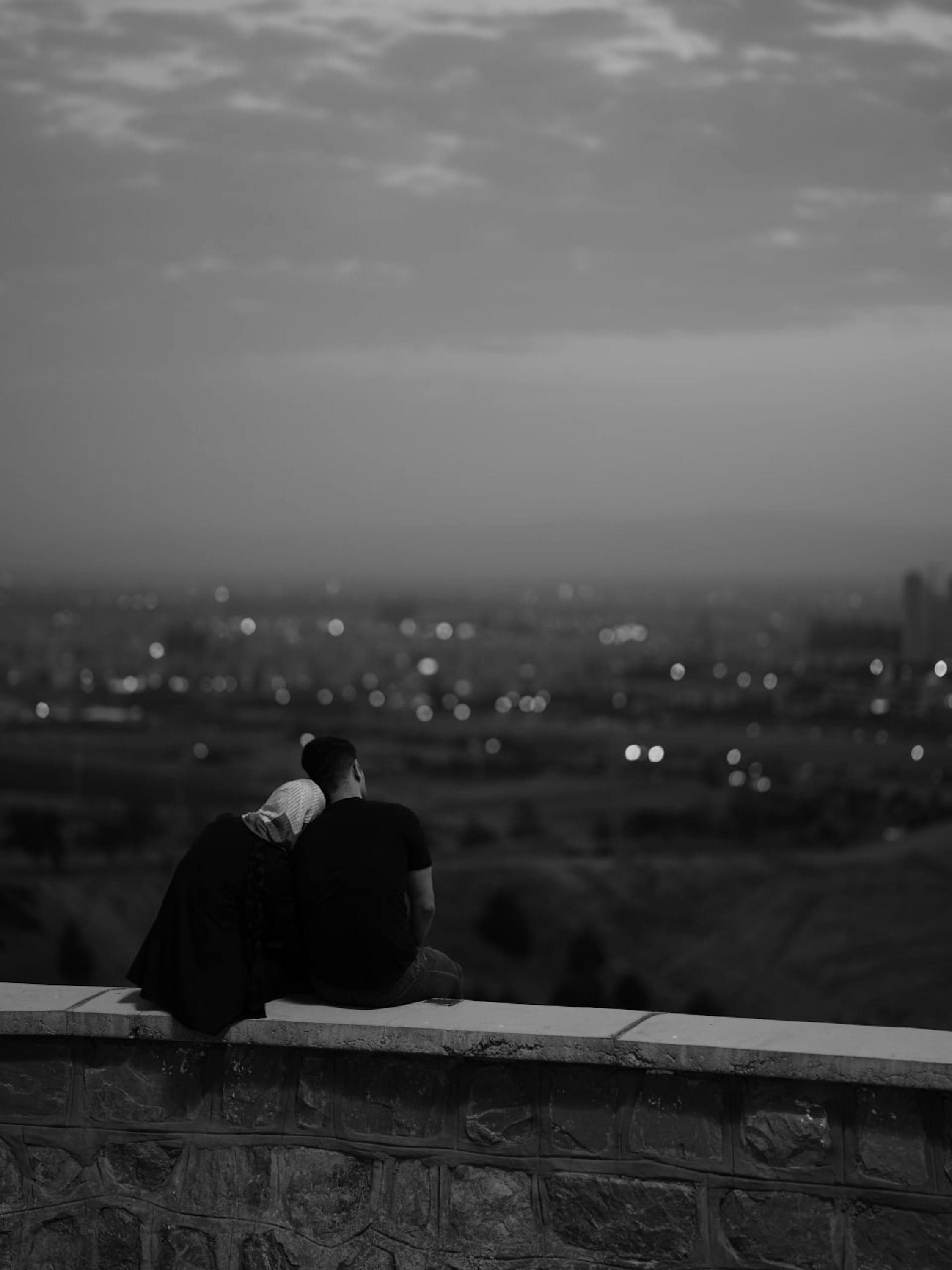 Black and white photo of a couple sitting on a wall, gazing at the city's lights at night.