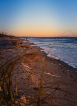 Person walks alone on a tranquil beach at sunset with waves gently lapping the shore.