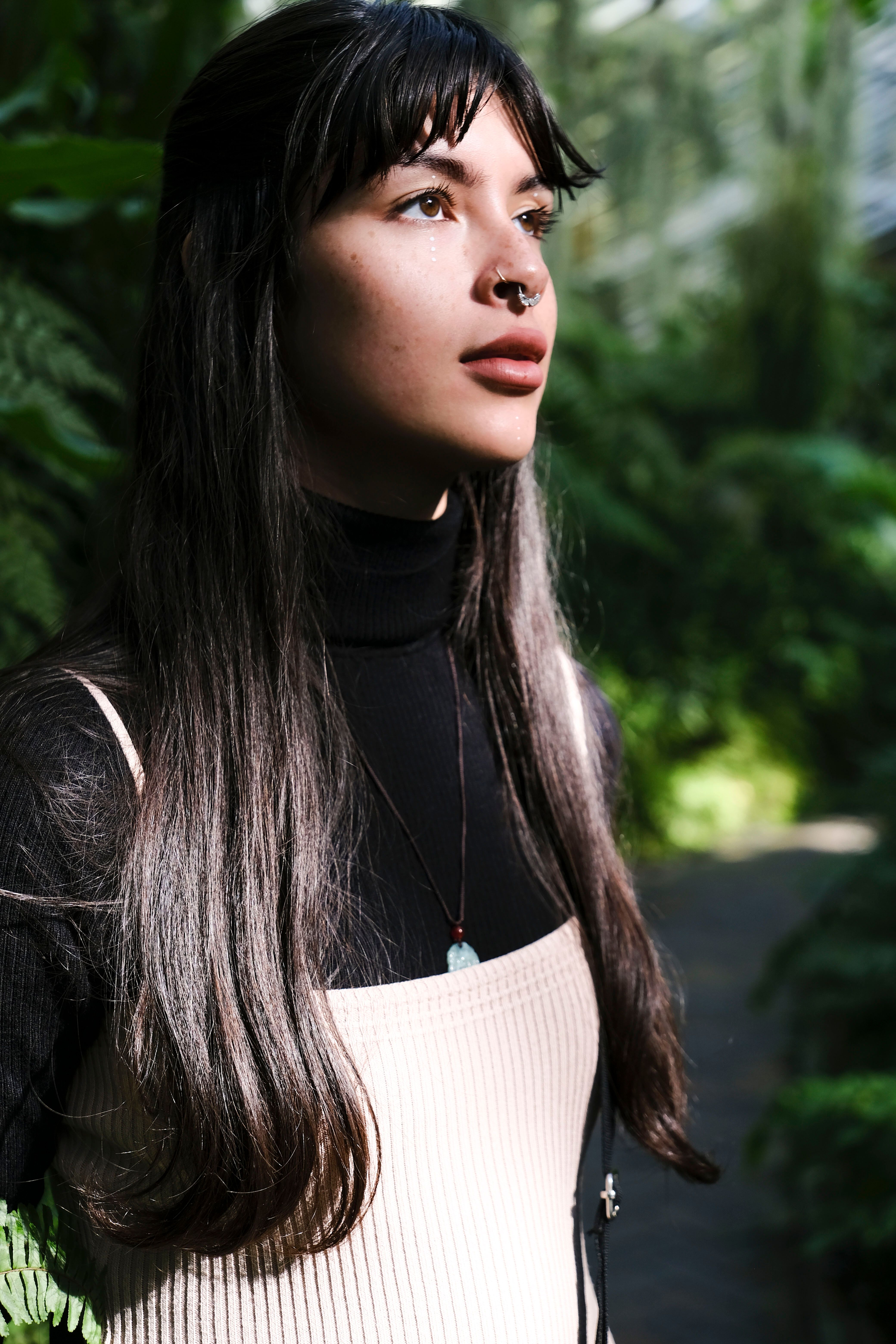A woman with long hair and a septum piercing stands amidst lush greenery, gazing thoughtfully.