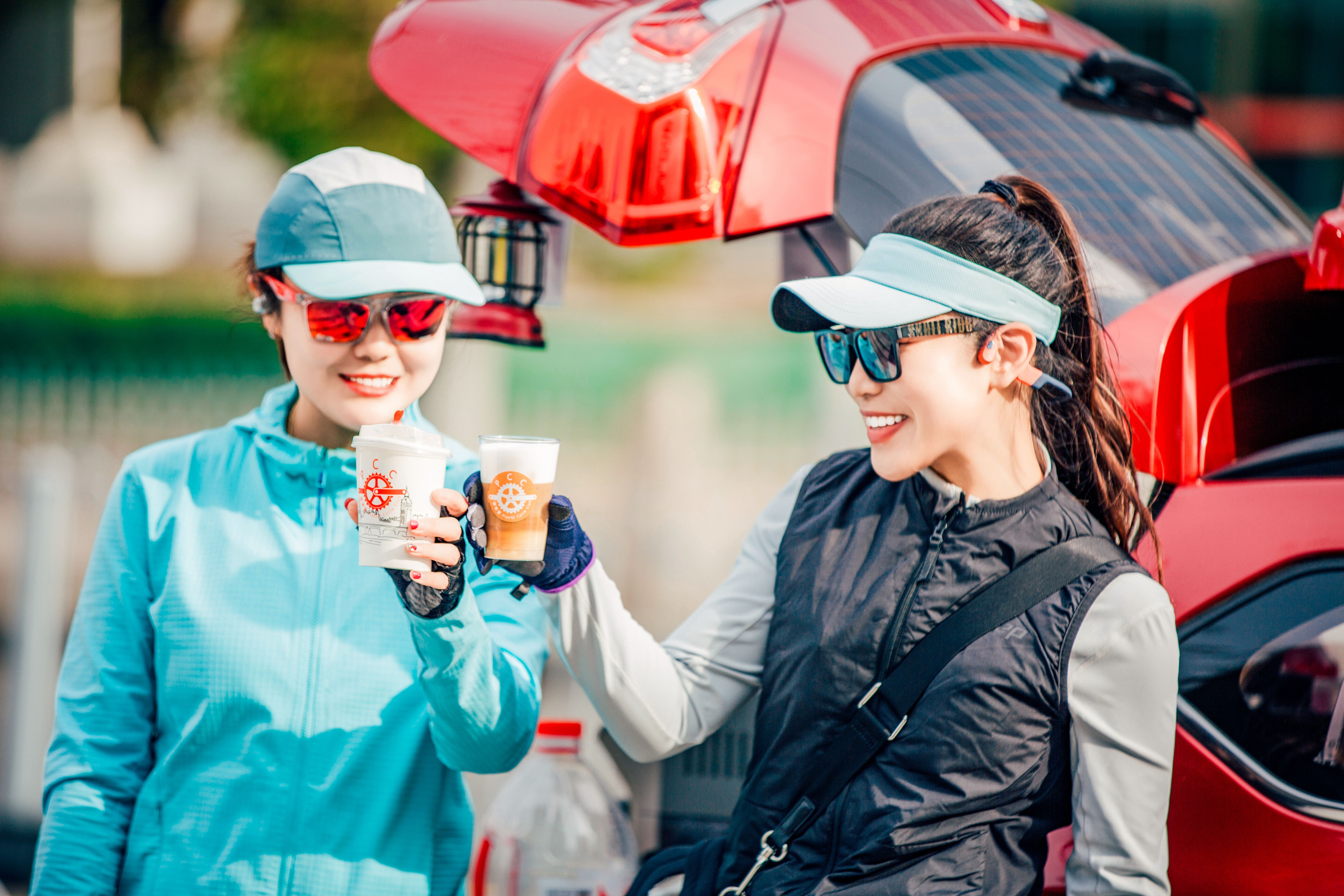 Two women enjoy cold drinks by a car trunk on a sunny day, smiling and wearing sporty sunglasses.