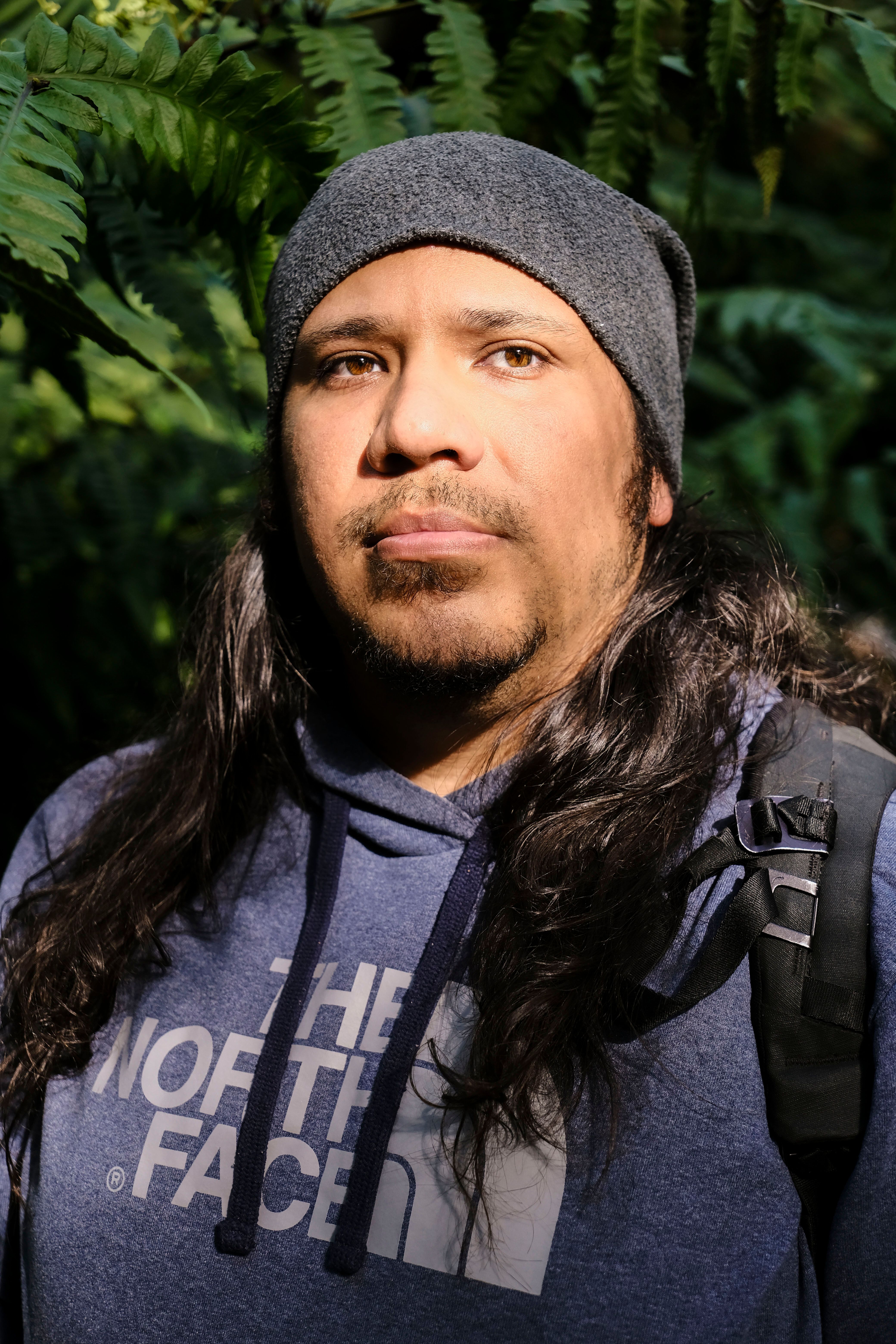 Casual portrait of a man in a beanie and hoodie standing among lush forest foliage.