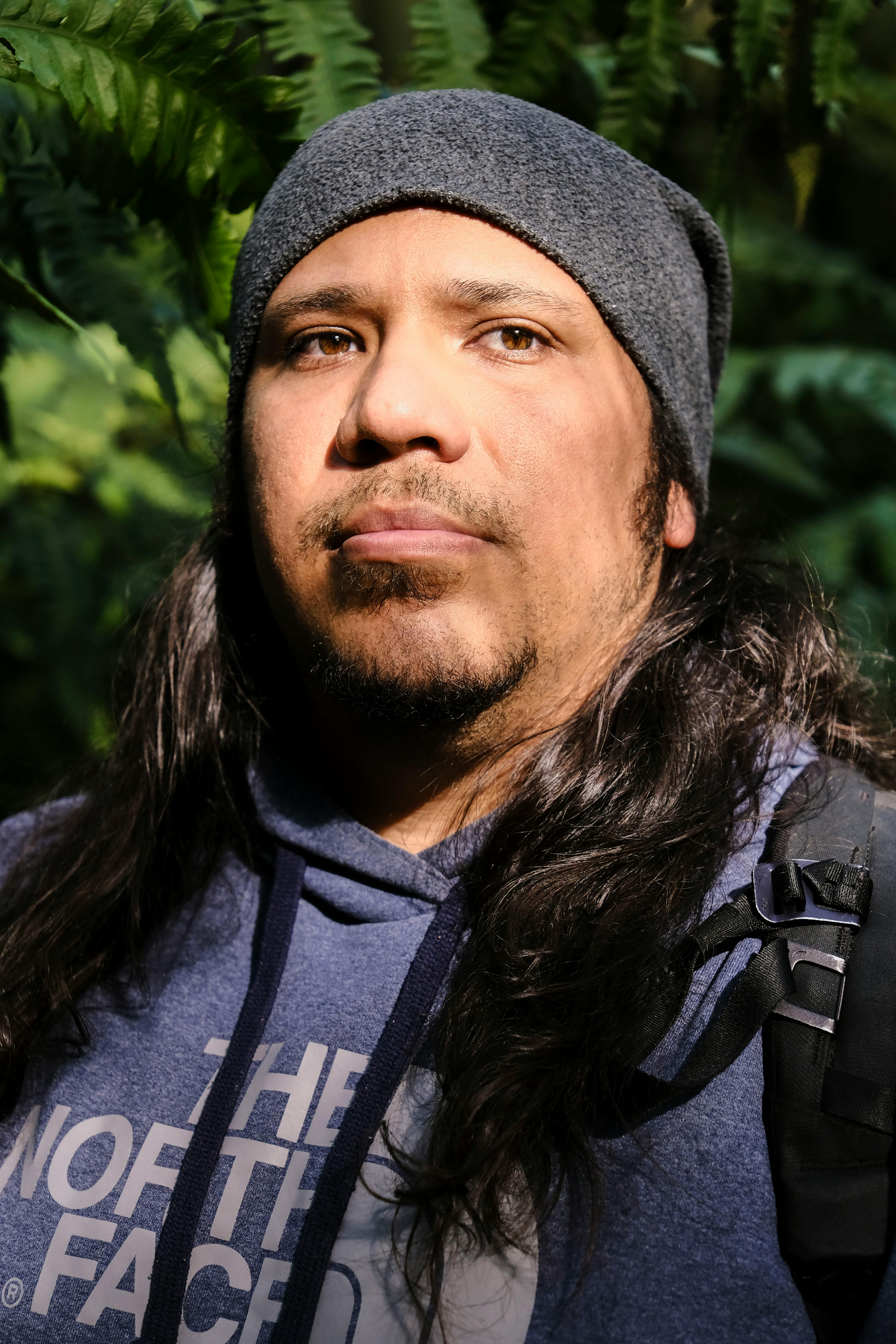 A man wearing a gray beanie stands outdoors surrounded by lush greenery, under natural light.