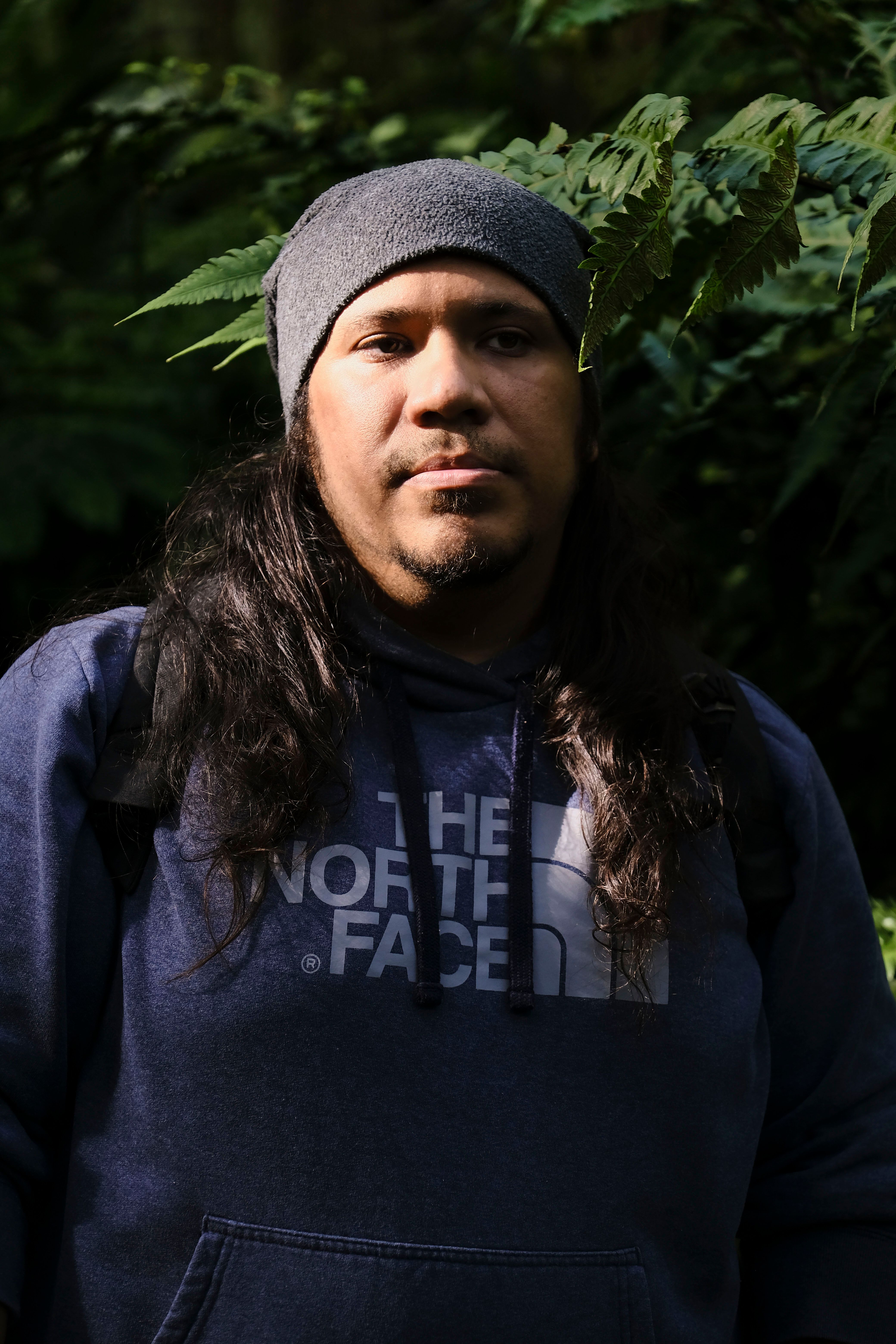 Portrait of a man in a blue hoodie and beanie surrounded by greenery outdoors.