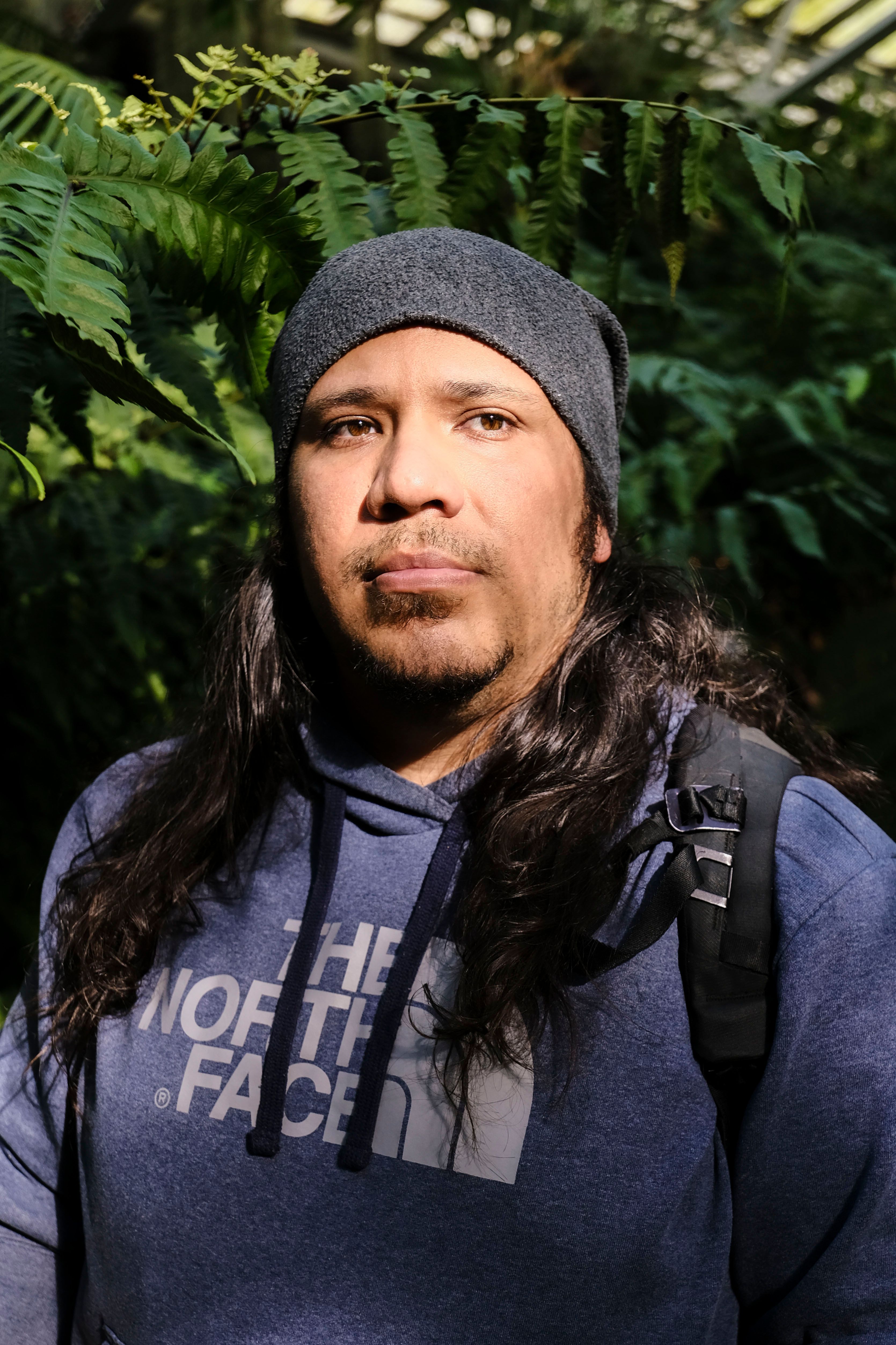 A man in a hoodie stands amidst lush greenery, sunlight highlighting his face.
