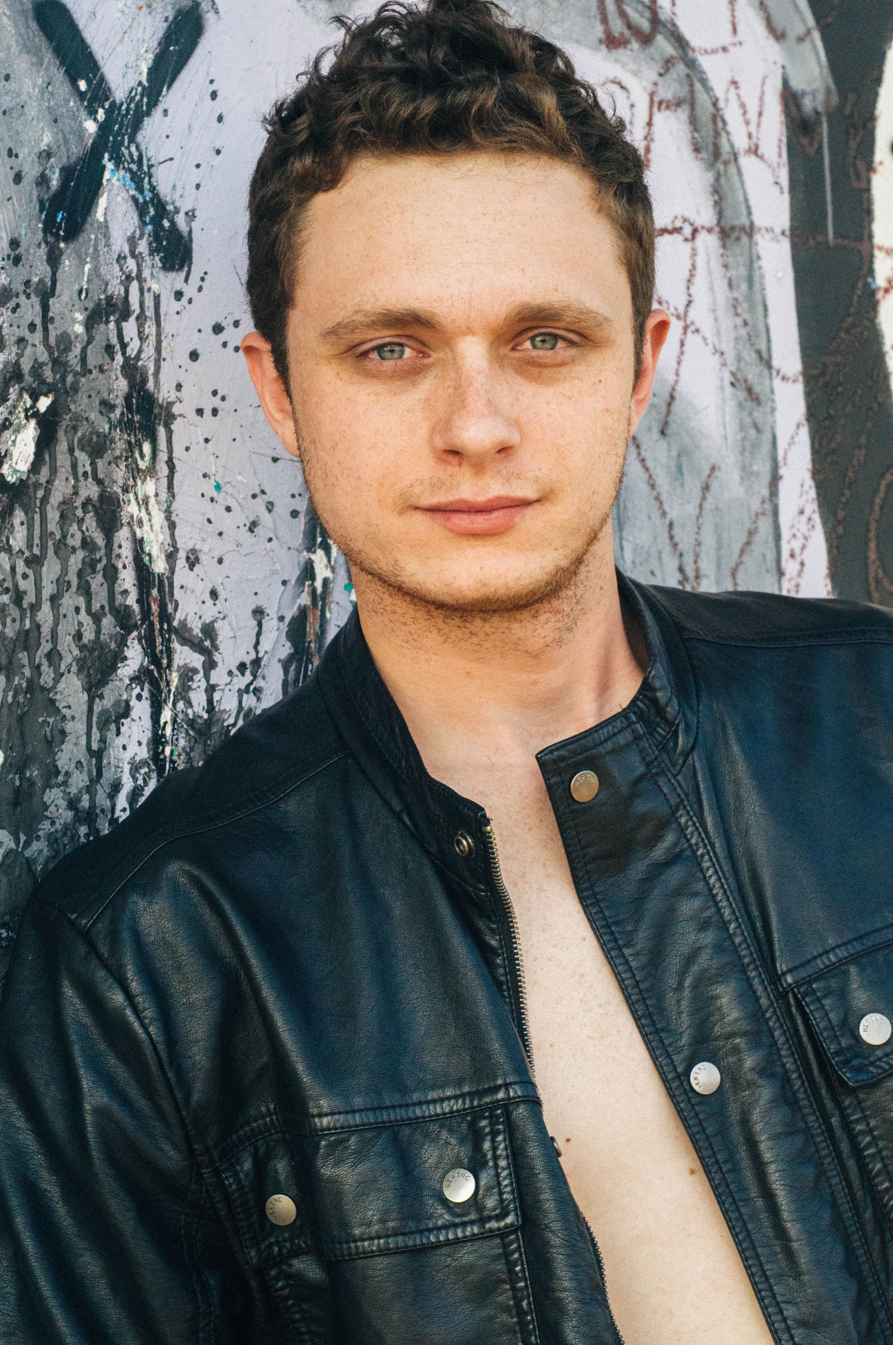 A young man poses casually in a leather jacket against a graffiti wall, expressing urban style.