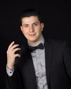 Sophisticated man in a black suit holding his bowtie against a dark background.