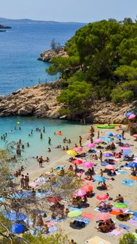 A lively beach scene with colorful umbrellas and numerous people enjoying the summer sun and blue water.
