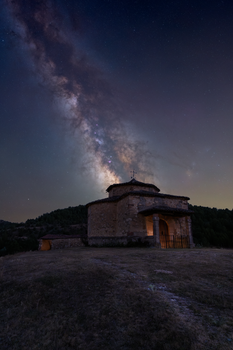 A breathtaking view of the Milky Way over a rustic chapel surrounded by nature under the night sky.