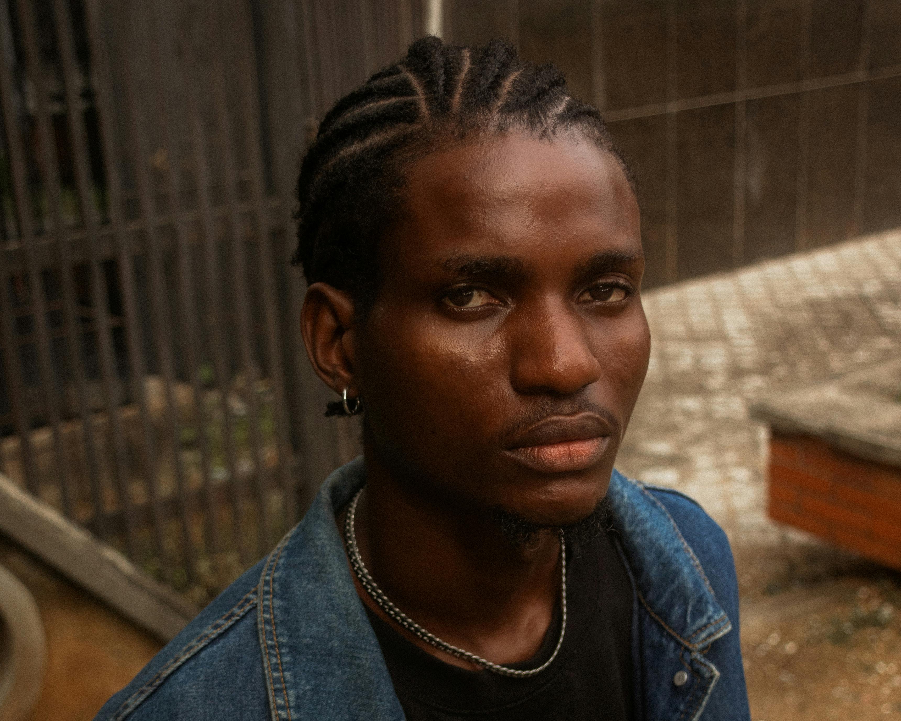 Close-up portrait of a young man with braids, wearing a denim jacket outdoors.