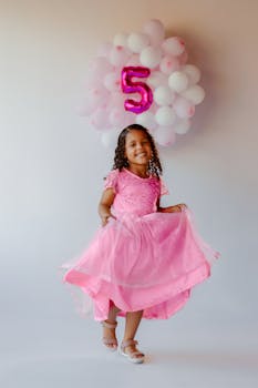 Smiling girl in a pink dress celebrating her fifth birthday with balloons.