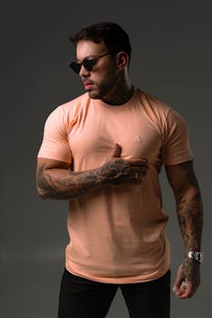 Confident man with tattoos in sunglasses posing fashionably in a peach t-shirt.