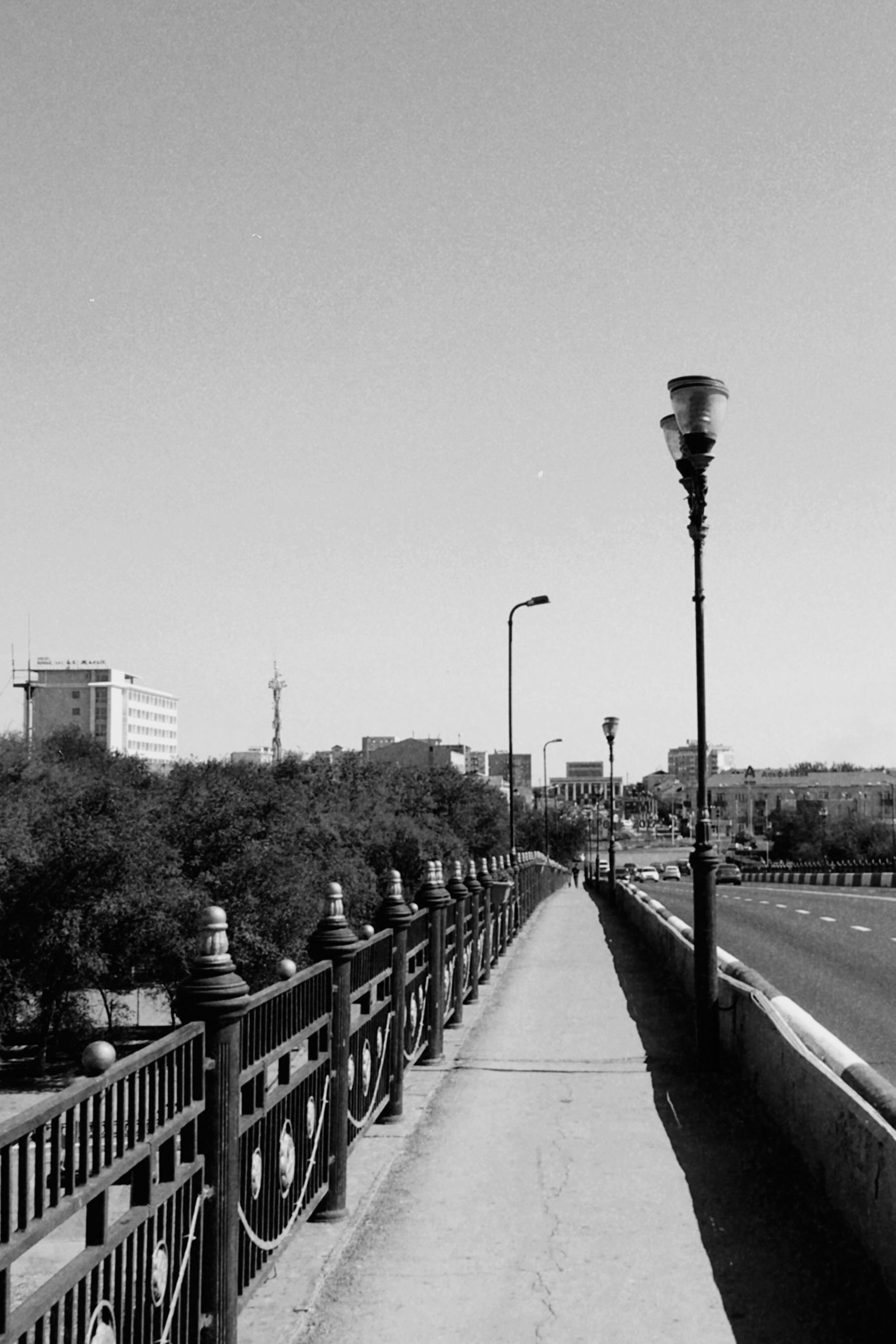 A serene black and white image capturing a walkway and road in Atyrau, Kazakhstan, with distant urban buildings.