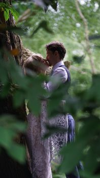 Romantic couple shares a loving kiss in a lush forest, captured through leaves.