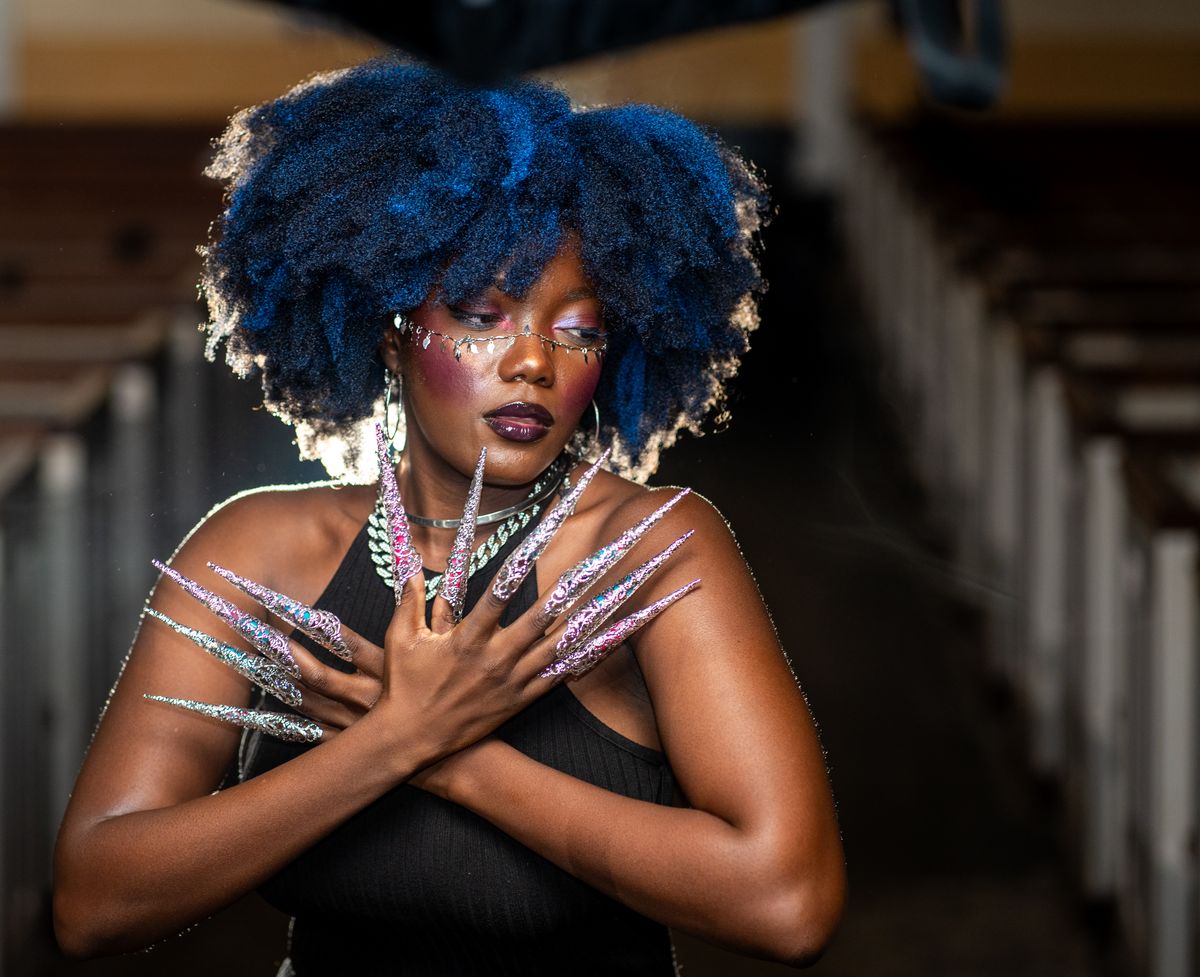 A striking portrait of a woman with blue afro and intricate nails poses creatively indoors.