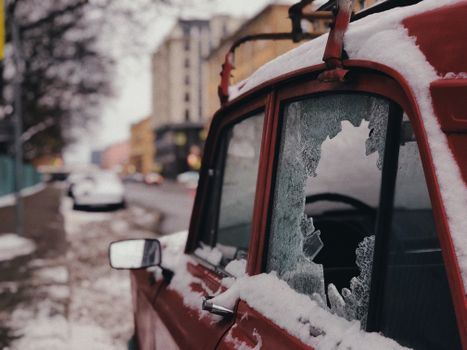 一辆红色汽车停在白雪皑皑的城市街道上，车窗破碎，凸显了冬季天气的影响