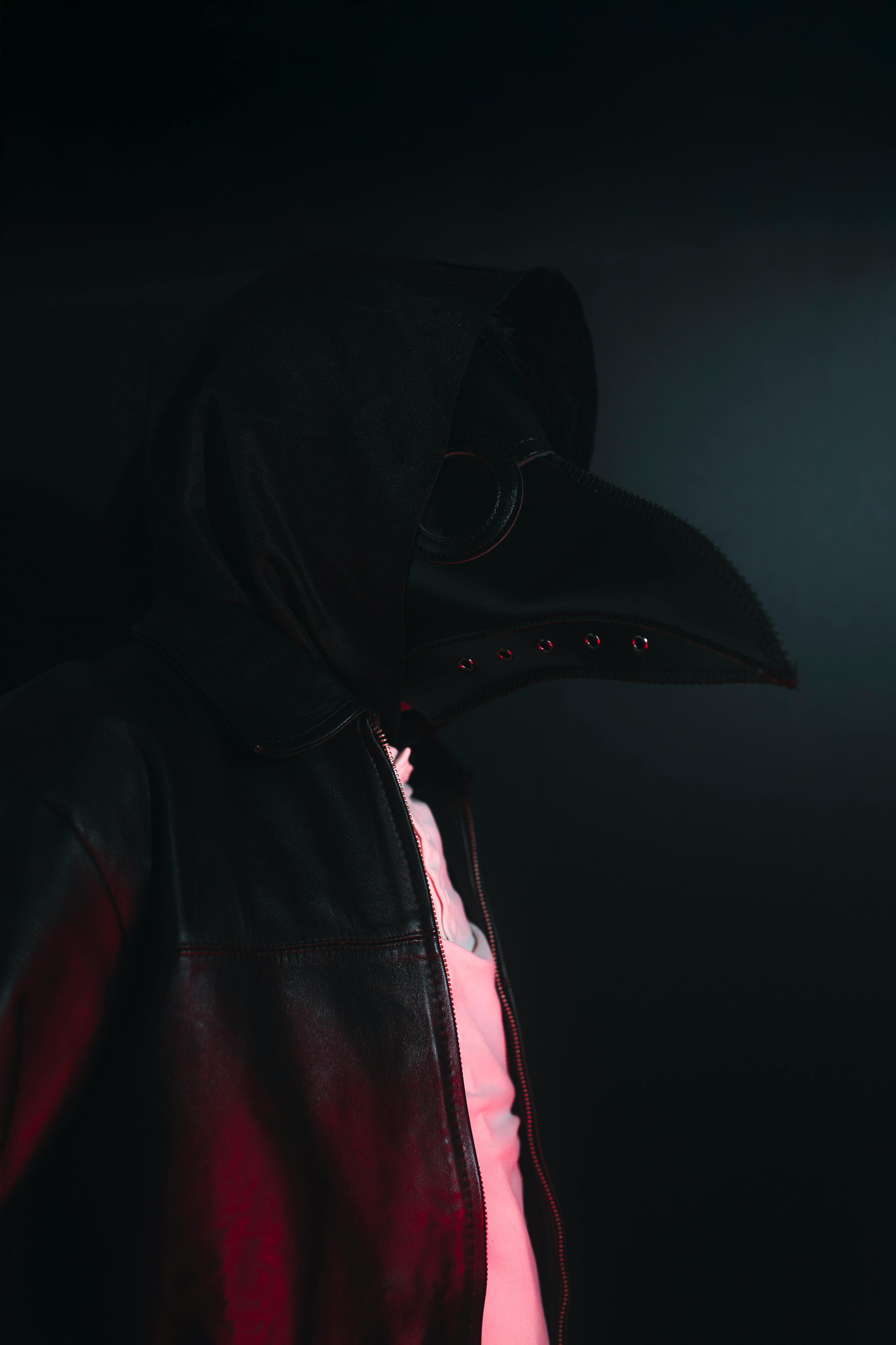 Gratis Una persona con un costume scuro da medico della peste e cappuccio, illuminata in modo spettacolare. Foto a disposizione