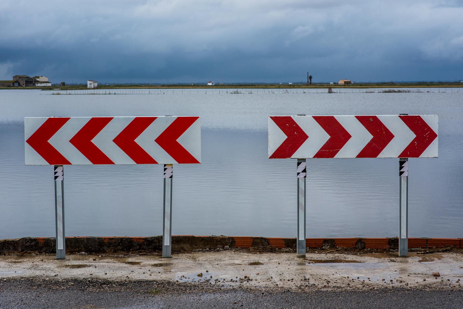 Red and white chevron road signs by a flooded area under cloudy skies