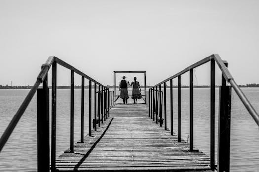 Two adults in traditional attire walk hand in hand on a wooden pier over still waters.