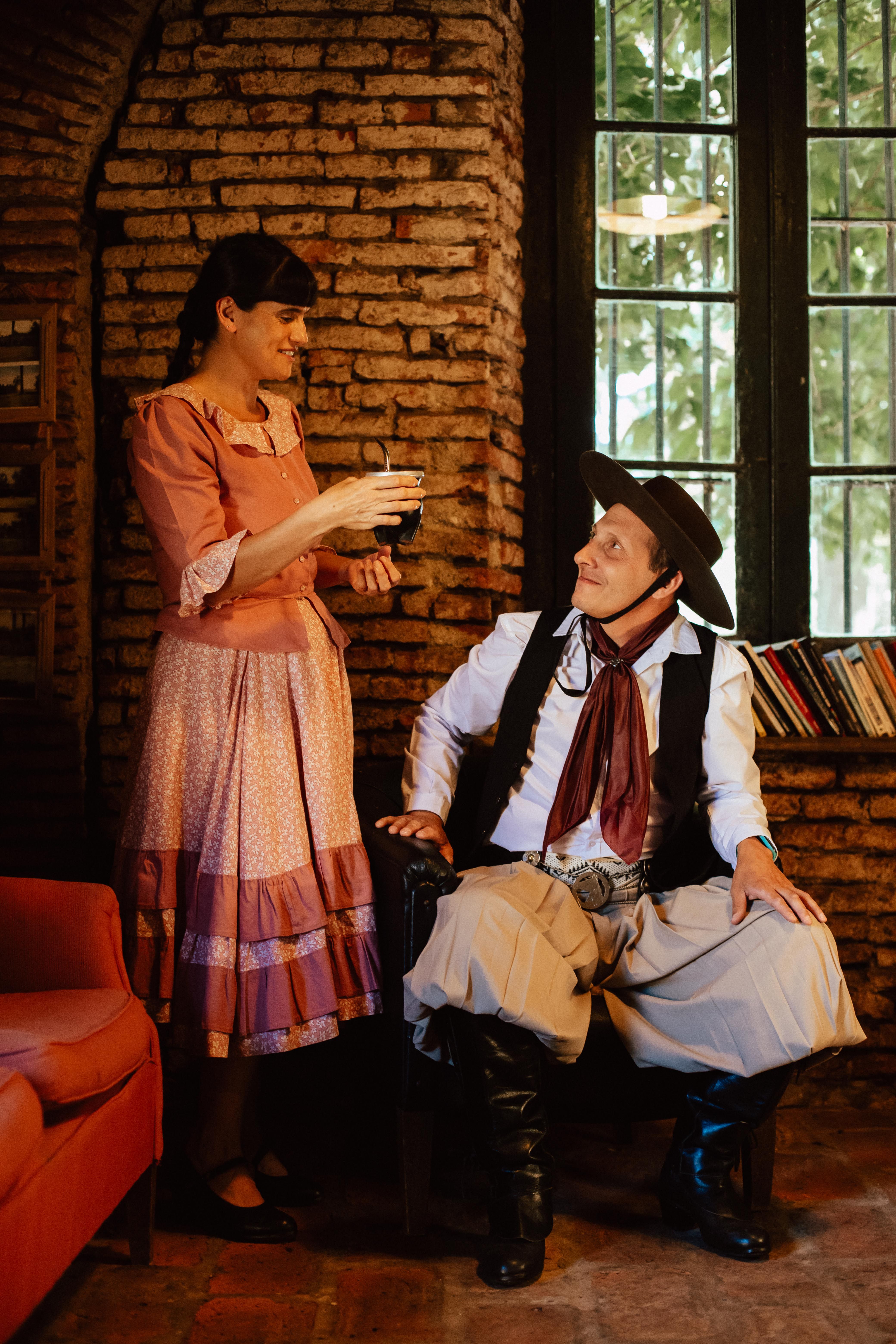 Argentine gaucho in traditional attire seated indoors with a woman offering a drink.